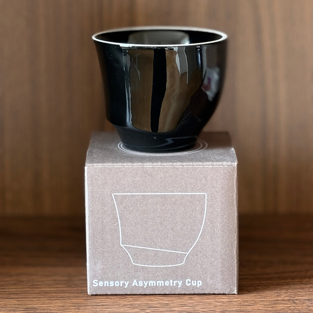 A glossy black sensory asymmetry cup displayed on top of its brown cardboard packaging box, set against a warm wood-grain background.