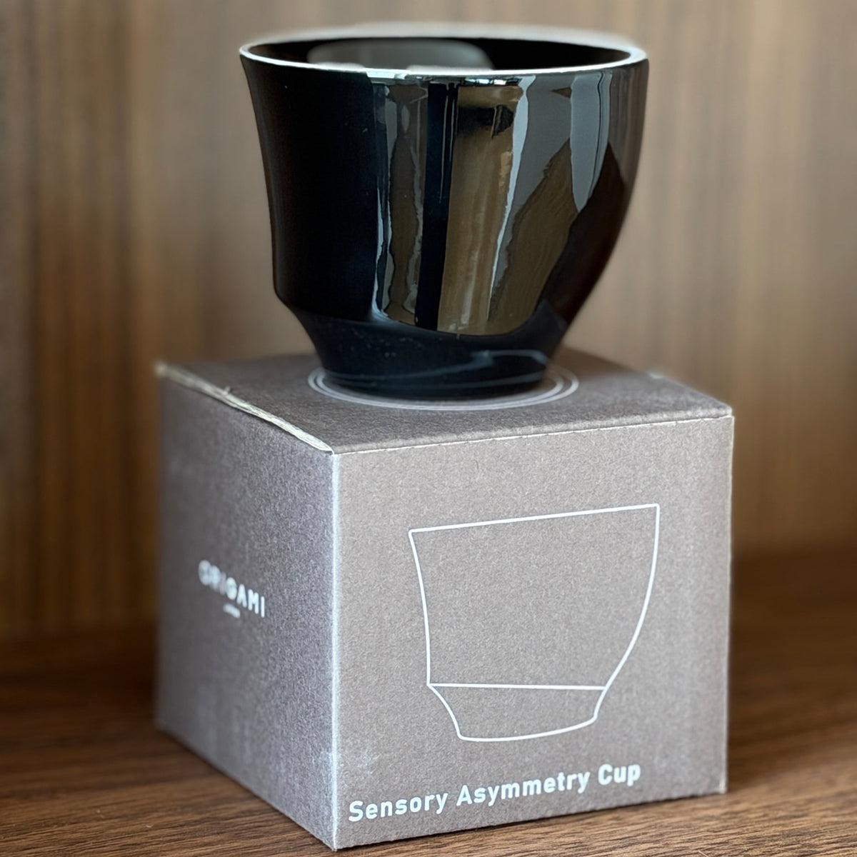 A glossy black Origami Sensory Asymmetry Cup displayed on its gray cardboard packaging box, set against a warm wood-grain background.